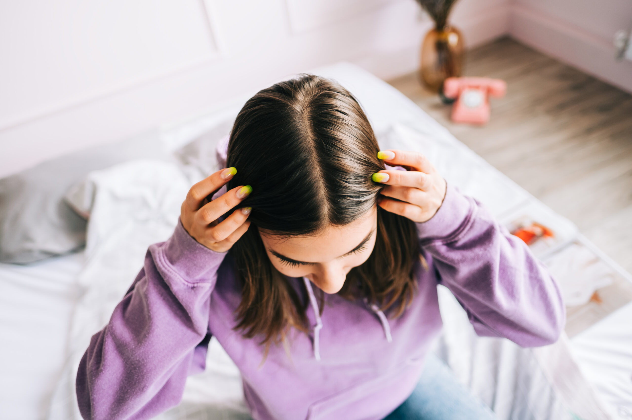 A woman with straight brown hair sits on the bed wearing a mauve top, and her hair looks healthy. haircare routine