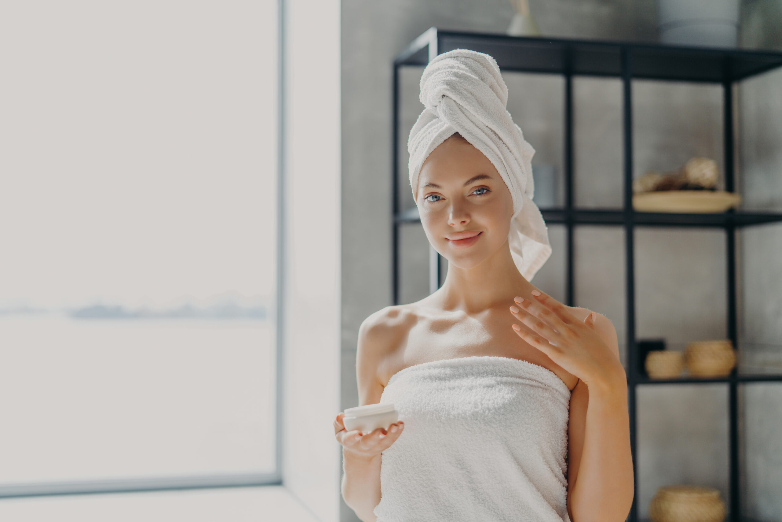 Elegant woman with a towel on her head holding a skincare product, waxes