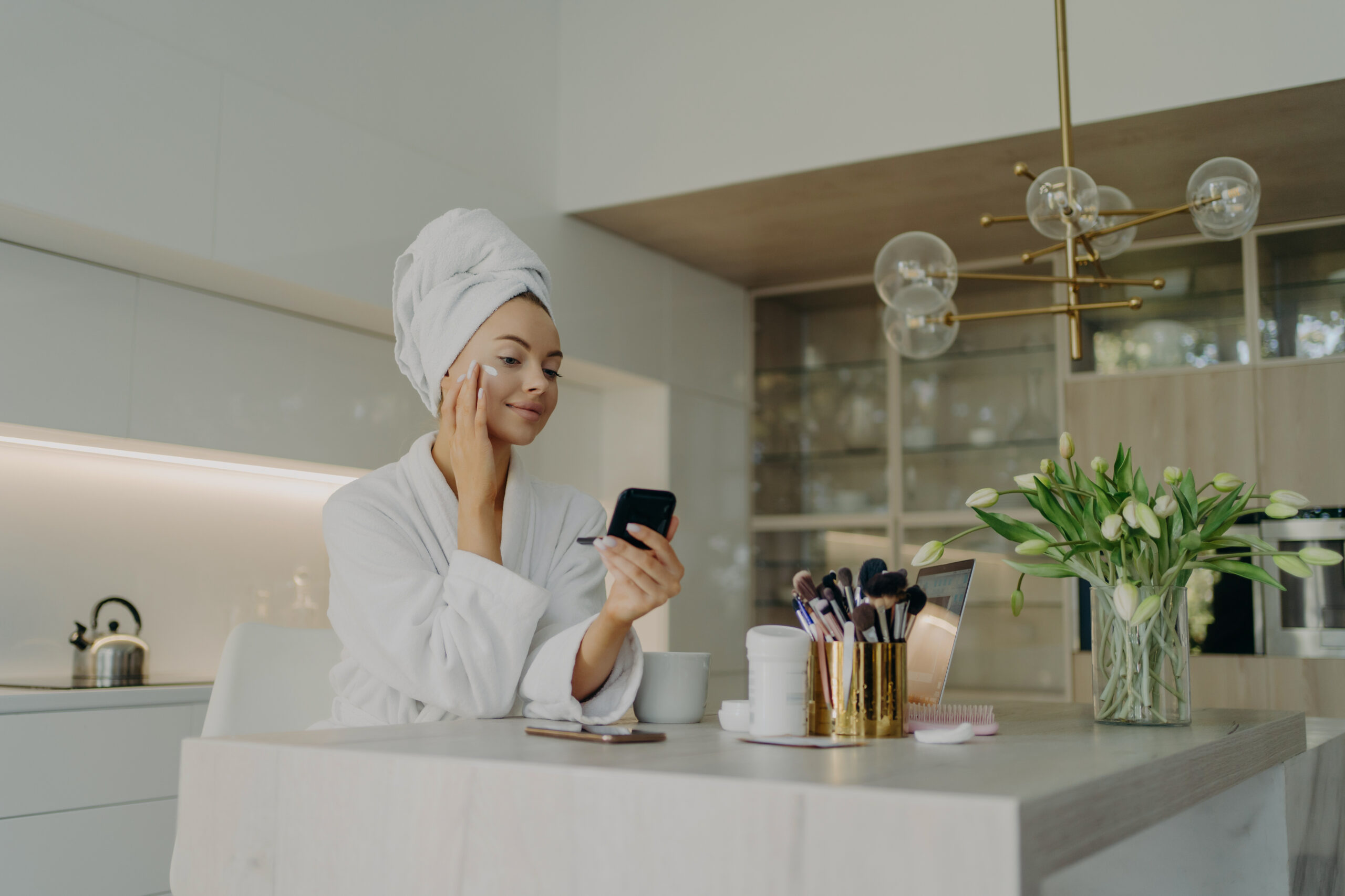 Beauty and facial care concept. Young pretty woman in white bathrobe applying cosmetic product on face and looking in compact mirror, taking care of her skin while sitting in modern kitchen at home