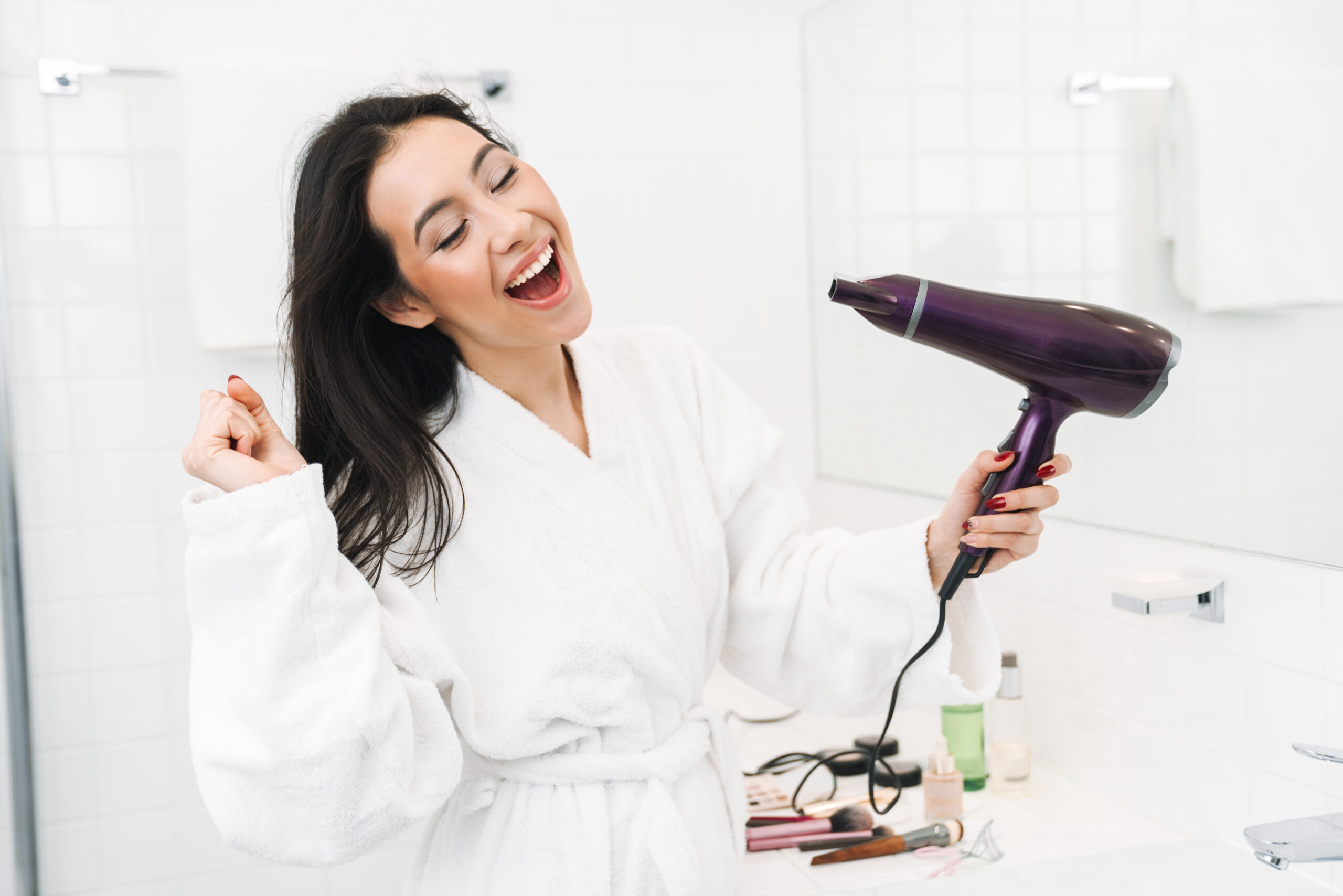 Image of a cute happy funny young woman indoors at home in bathroom dry hair with hair dryer singing. hair tools