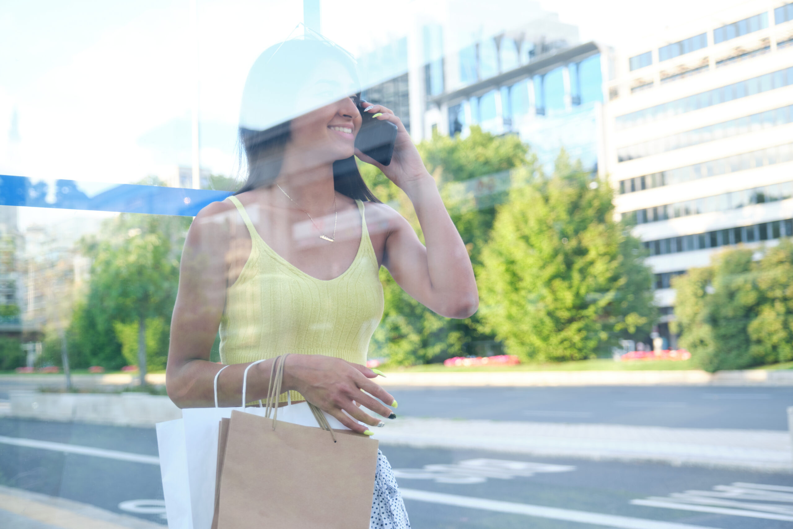 Young latin woman talking on the smartphone while is waiting for the bus at a bus stop, holding shopping bags at street.