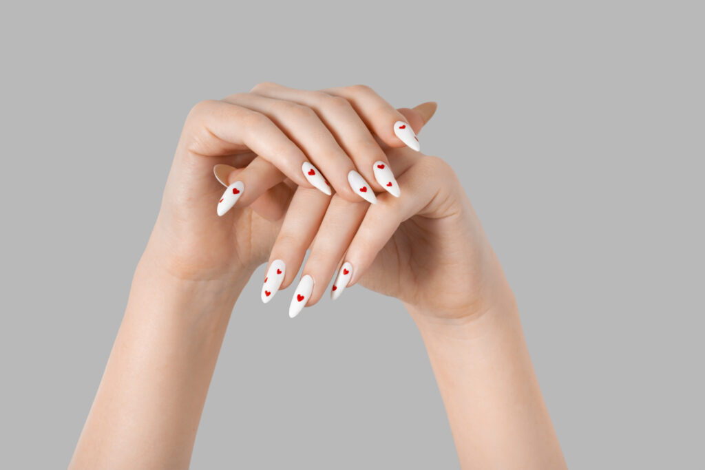 Stiletto Nails. Female manicure two hands minimal gel polish white long nails and red hearts desing gray isolated background. Beauty spa, Valentine's Day, love concept.
