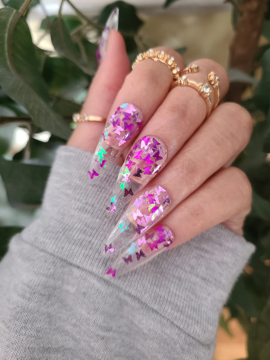 A woman's nails, wearing gold rings on her fingers with encapsulated flowers painted in purple