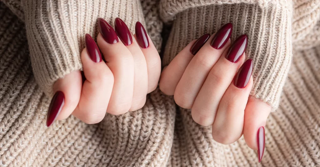 A faceless woman showcases stiletto nails adorned with a deep red nail shade, complementing her attire of a light brown sweater.