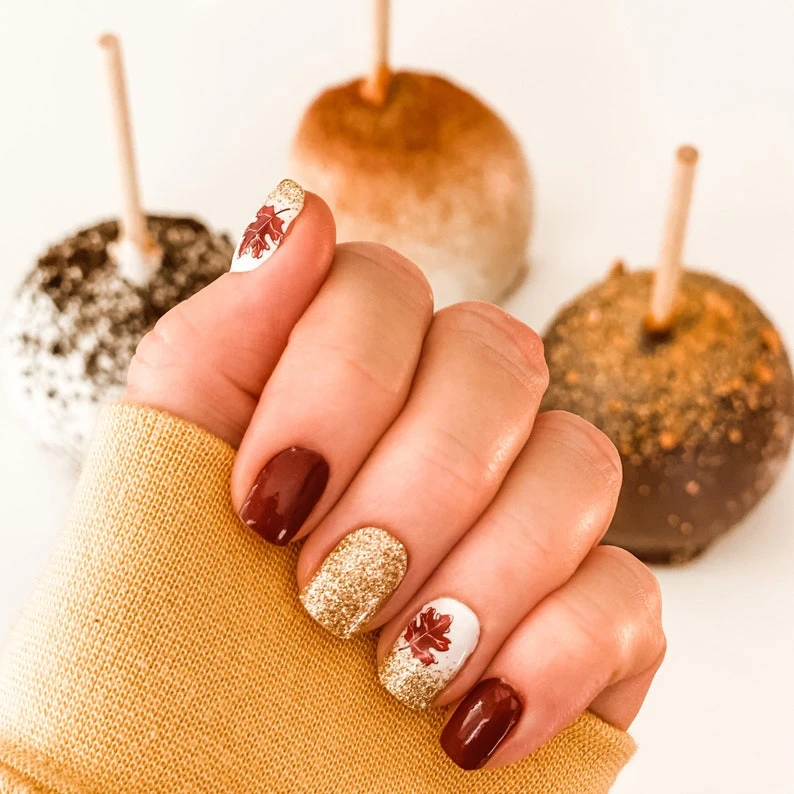 A woman's nails with a design of fallen autumn leaves on her nails, wearing a fall yellow sweater