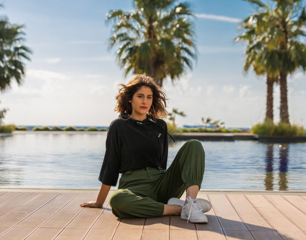 Nour, who is influential in the world of beauty, wears a black sweater and olive-colored pants, with a lake behind her