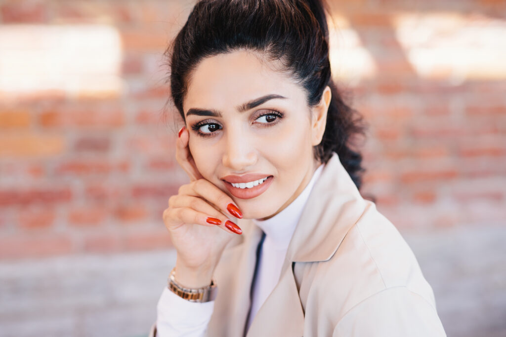 Close up of glamour brunette female having thin eyebrows, dark eyes with long eyelashes, pure skin, full well-shaped lips, red manicure wearing elegant watch on hand and white jacket looking aside. everyday manicure