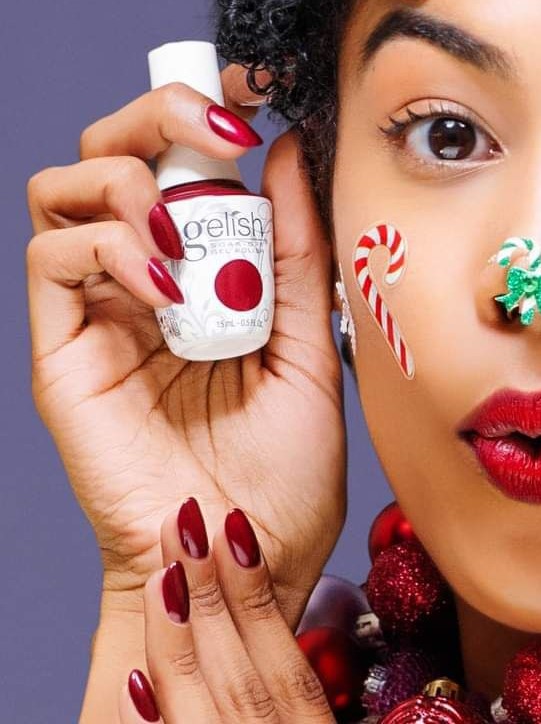 A woman holding a bottle of Gelish On My Wish List nail polish with Christmas decorations in the background.