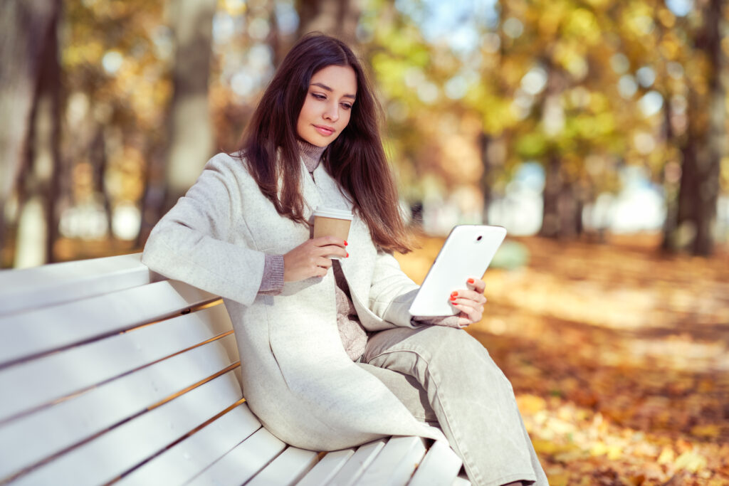 Young beauty woman sitting with a cup of coffee on a bench in the fall park, her nails painted in red nail shade. A girl reads an e-book in the autumn forest