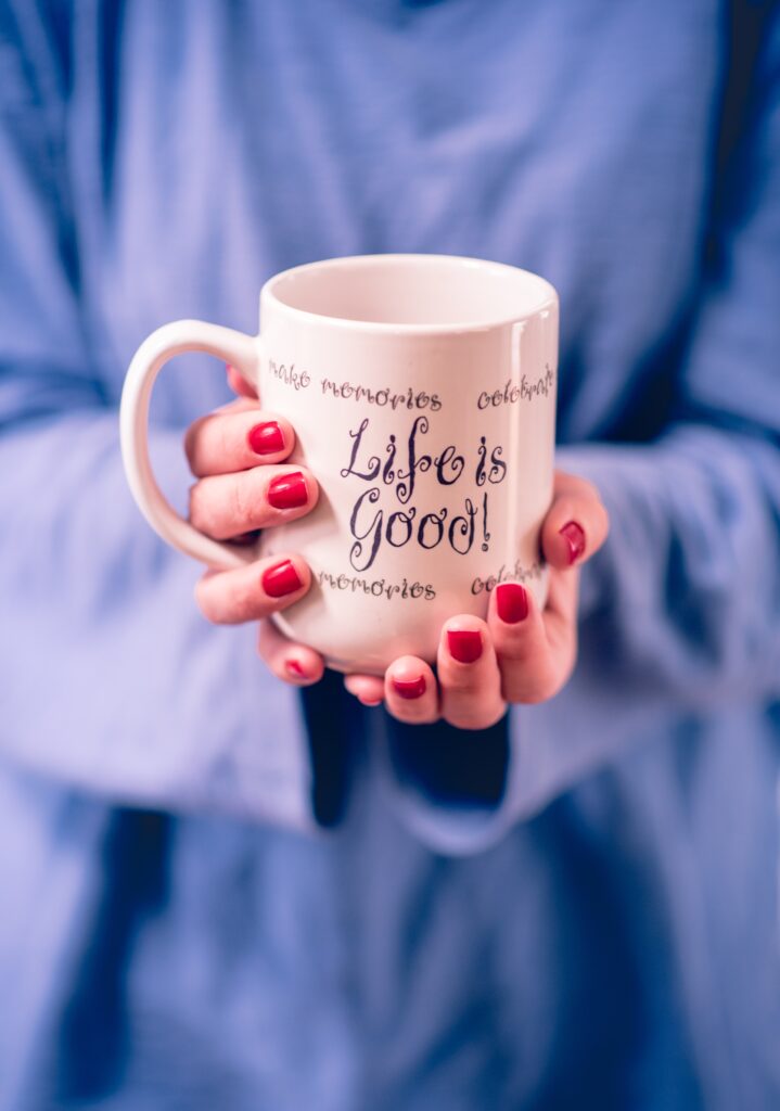 young millennial woman at home in blue shirt enjoying a glass of coffee from mug red fingernails Red nails. Red manicuer