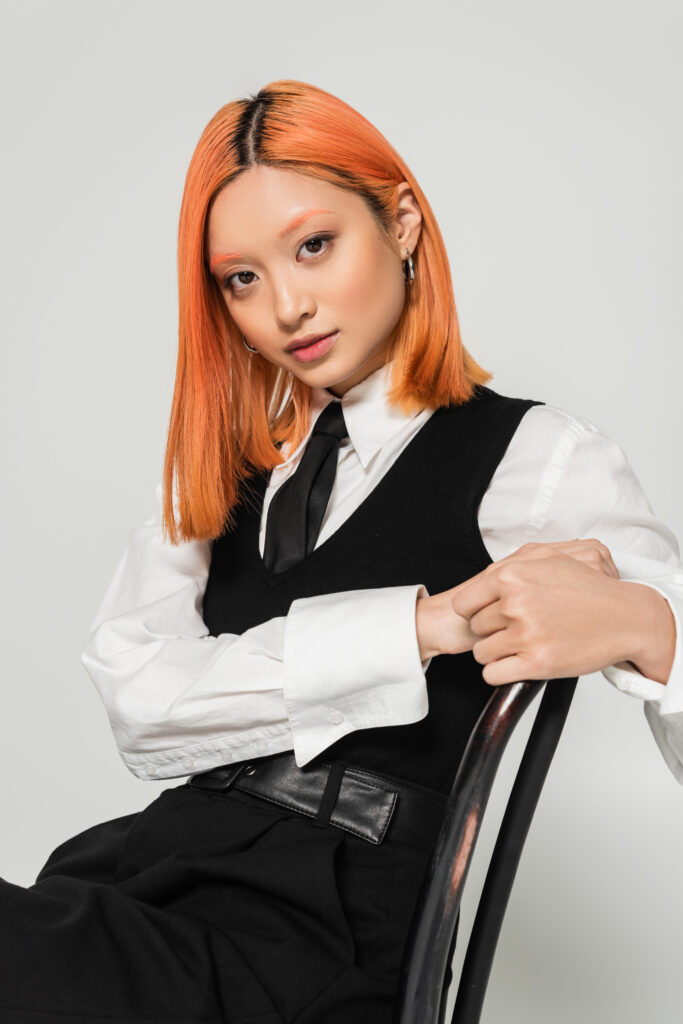 Appealing and young Asian woman with dyed red hair, wearing white shirt, black vest and tie, sitting. Dyed hair healthy
