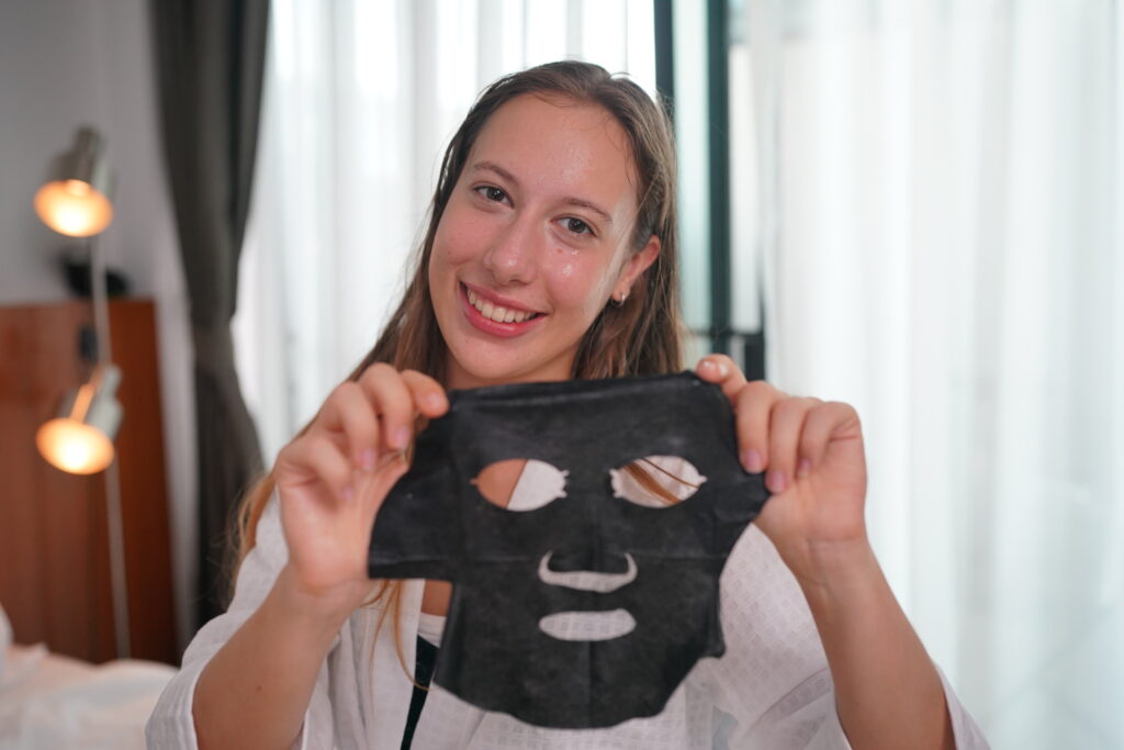 Young woman applying facial masks. She's sitting on her bed looking at mirror and taking care of here skin during weekend morning.