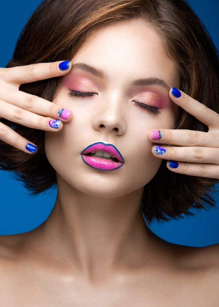 Beautiful model girl with bright makeup and colored nail polish. Beauty face. Short colorful nails. Picture taken in the studio on a blue background.