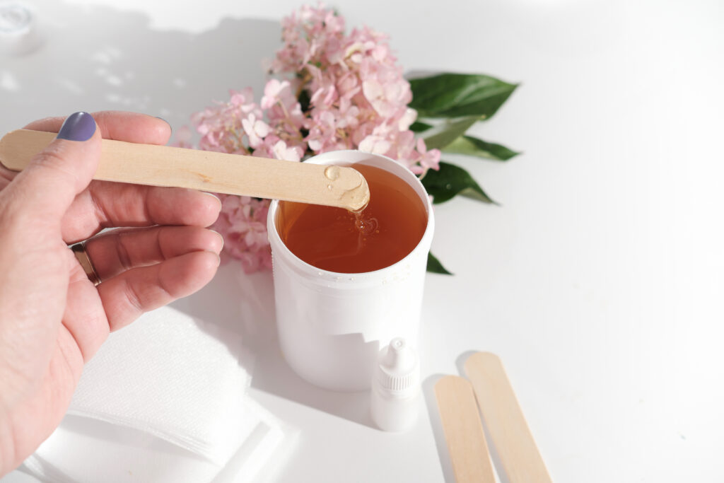 female hand holding a spatula with soft wax paste over white plastic container. depilation set next to pink flowers on a white background. beauty and skincare concept.