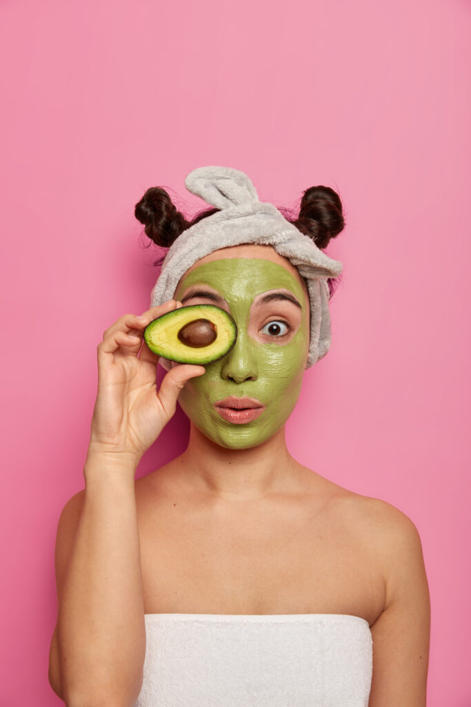 Natural cosmetics concept. Asian woman applies green nourishing mask on face, covers eyes with half of avocado, wears headband, has two buns, stands wrapped in towel, relxes during spa treatments