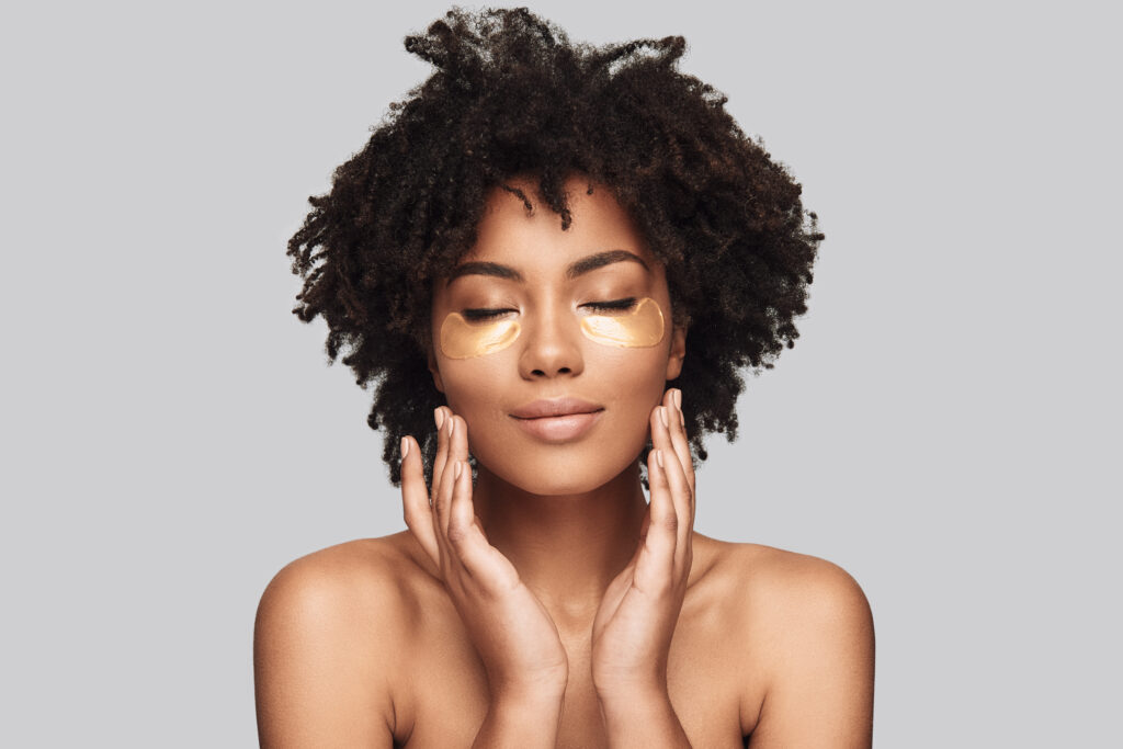 skin care. Attractive young African woman applying medical eye patch and smiling while standing against grey background