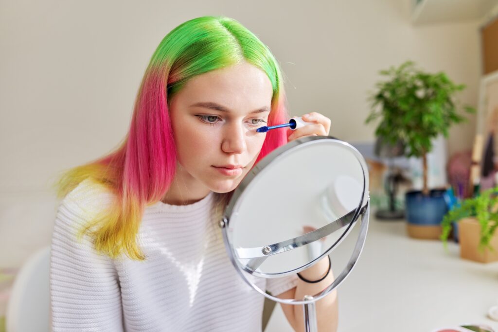 Cosmetics and makeup for teenagers, fashionable girl with colored dyed hair dyeing eyelashes with mascara looking in the makeup mirror at home near the window, Haircut and Color Trends