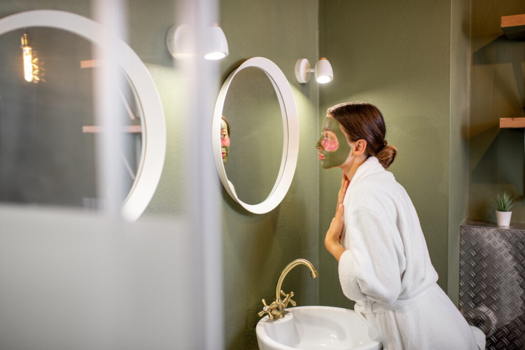 Woman in bathrobe taking care of herself with facial masks looking into the mirror in the beautiful green bathroom