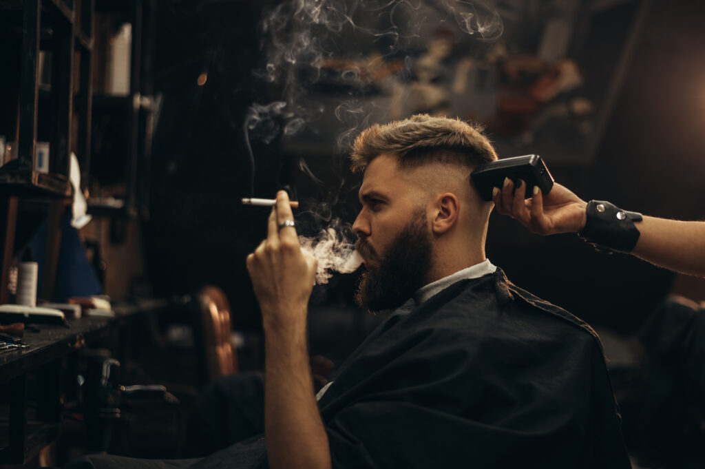 Young bearded man getting haircut by hairdresser while sitting in chair at barbershop and smoking a cigarette