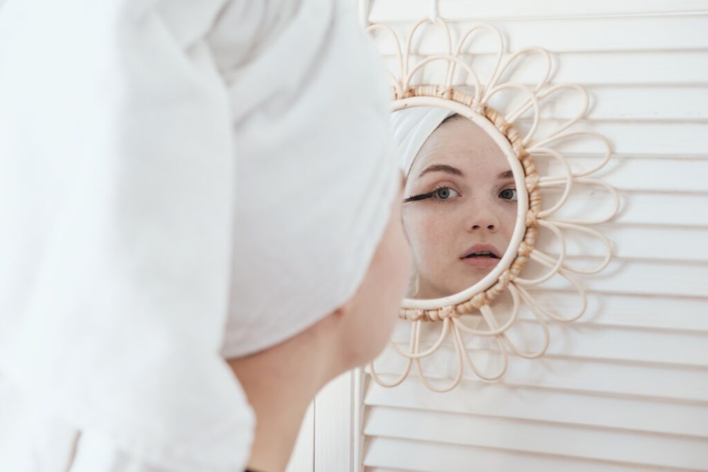 A young beautiful woman with a towel on her head after a shower in the bathroom paints her eyelashes, looking at her reflection in the mirror on the wall. Concept of female morning makeup procedures.
