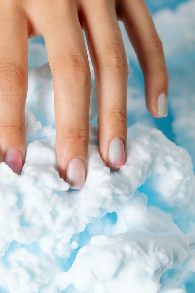 A close-up image of a hand with long and healthy nails on soft, fluffy clouds against a backdrop of a bright blue sky.
