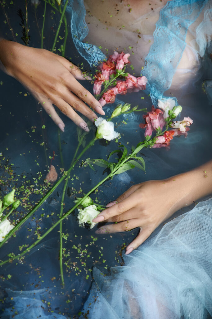 Art beautiful romantic woman lies in swamp in blue long dress with flowers. Portrait brunette in transparent dress in water swamp mud duckweed. Spring nail polish colors trends for 2024