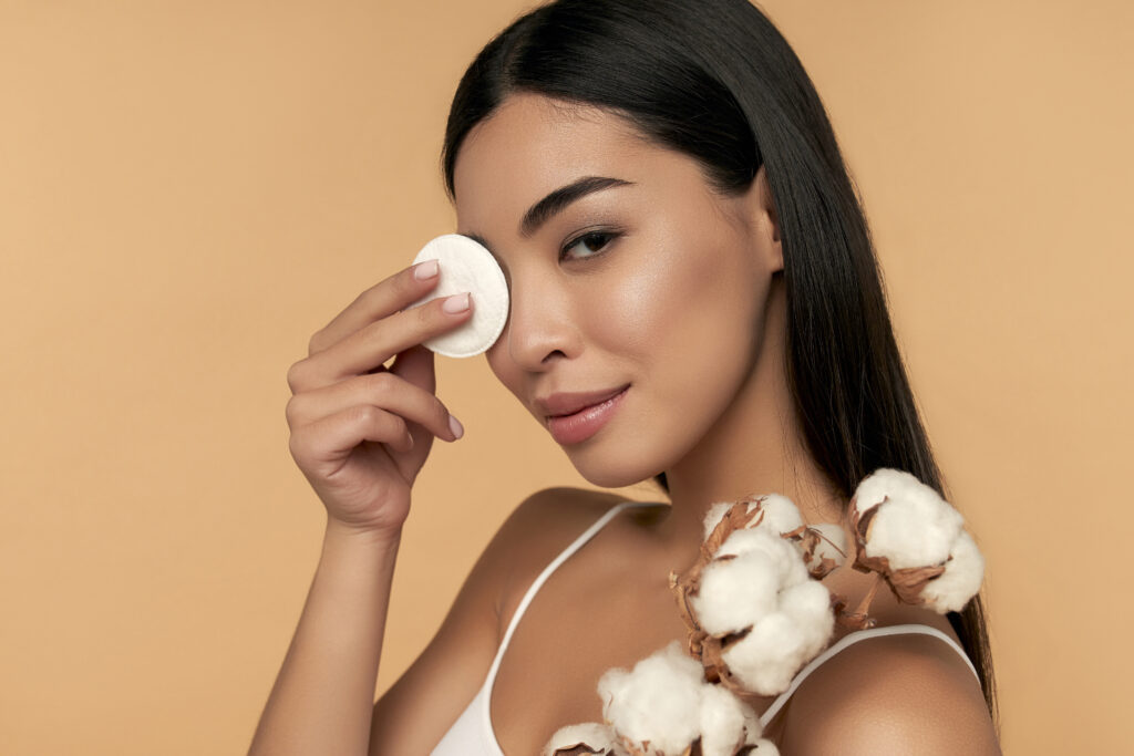 Young woman cleans beauty face using cotton pads, cleansing lotion and face toner to remove makeup. Nice girl with beautiful skin. Skin care concept. Beige background, removing eyeliner