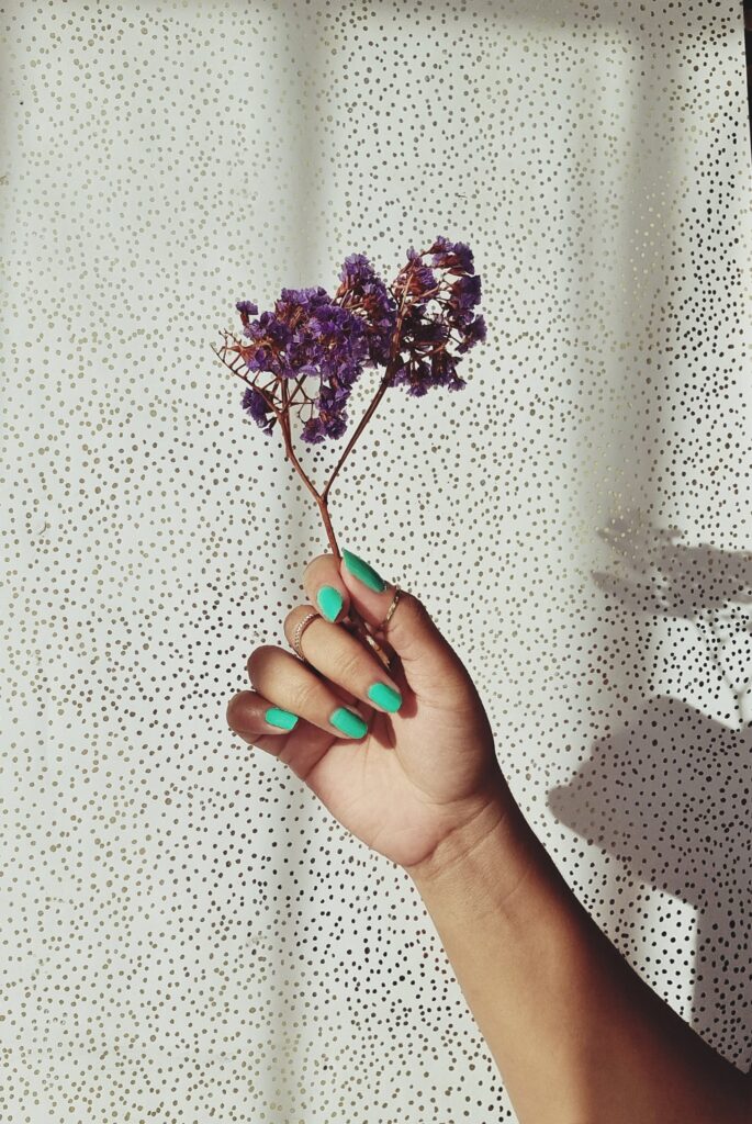 A woman's hand with light green painted nails holding a lavender plant against a white wall with black dots.