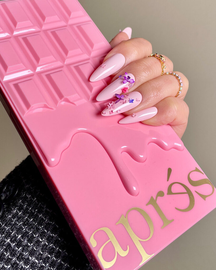 A faceless woman's hand is holding the Apres Gel-X nail tips kit. Her nails are painted in a pink nail polish shade with floral and butterfly nail designs.