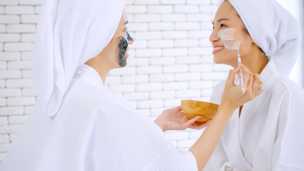 Beautiful young woman in white bathrobe applying a revitalizing white mask onto her friend's face. morning skincare. facial care, face care. facial masks
