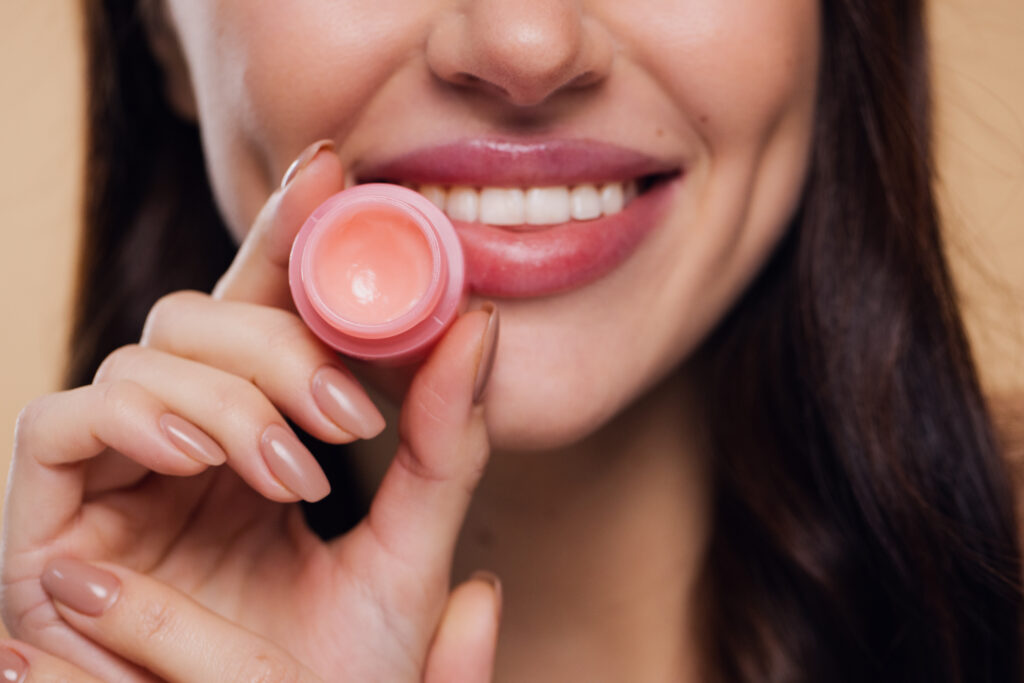 Close up beauty portrait of smiling woman cares of her lips on beige background. Young brunette woman applying make-up cosmetic SPA product. Model using lip balm or gloss for hydration, nutrition, smoothing balm. lip care