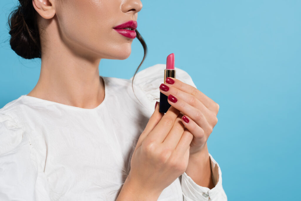 cropped view of young woman in white blouse holding pink color lipstick isolated on blue