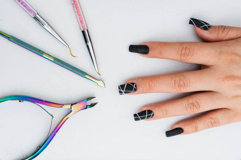 hand of a girl with her nails made in polygel on a white background and next to her the ideal elements for a basic manicure. cuticle nippers, cuticle pusher and fine painting and design brushes. 2024 nail trends