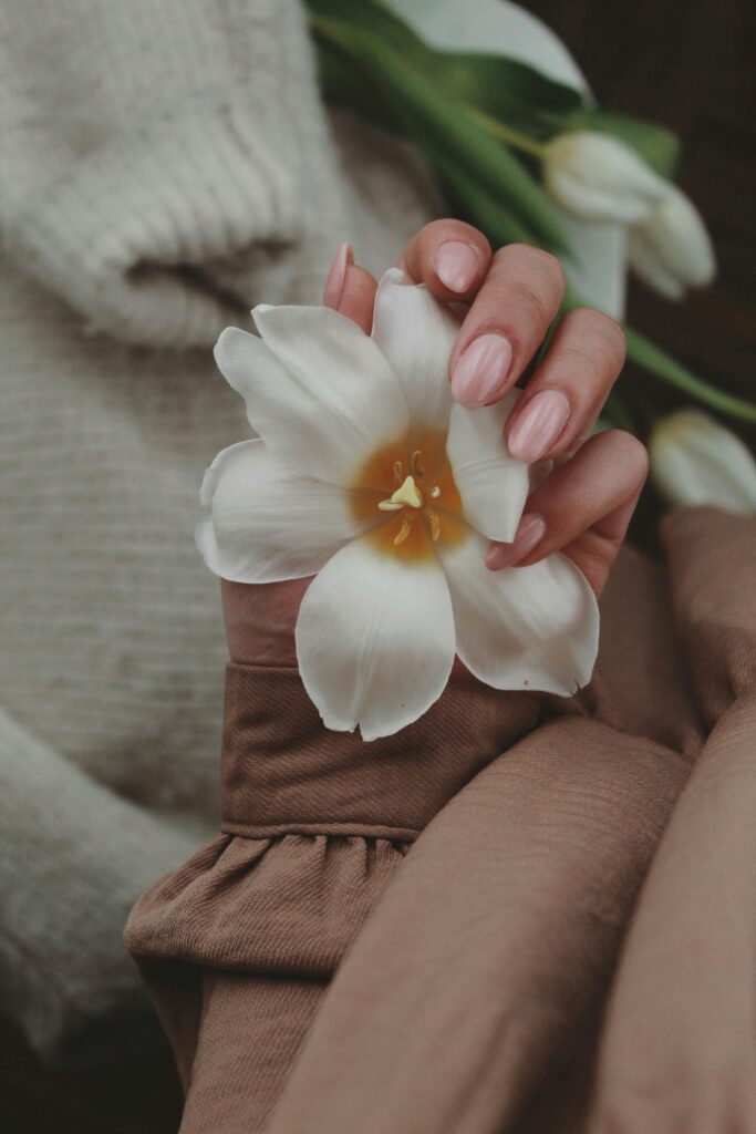 A faceless woman holding a white flower her nails are painted with light pink nail polish shade