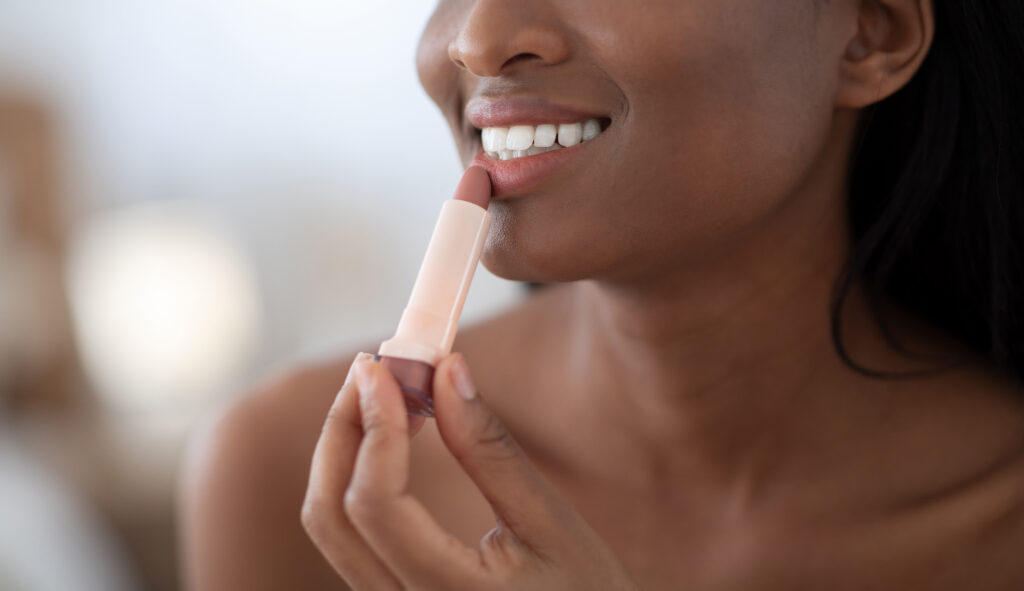 Smiling young beautiful african american lady applying nude color lipstick on lips get ready in morning. Woman look in mirror, doing make up put cosmetic on face in modern bedroom interior, cropped. Lip care