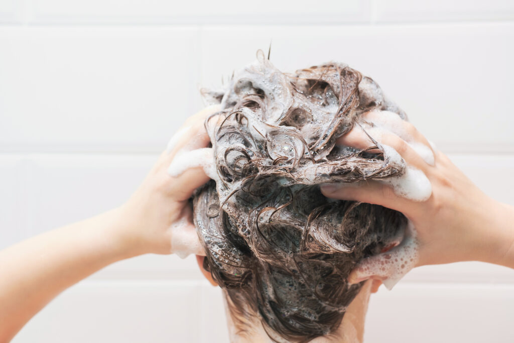 A young woman washes her hair with shampoo on a white tiles background, back view.