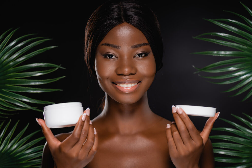 Day vs. Night face Moisturizers. African American woman with face cream near green palm leaves isolated on black