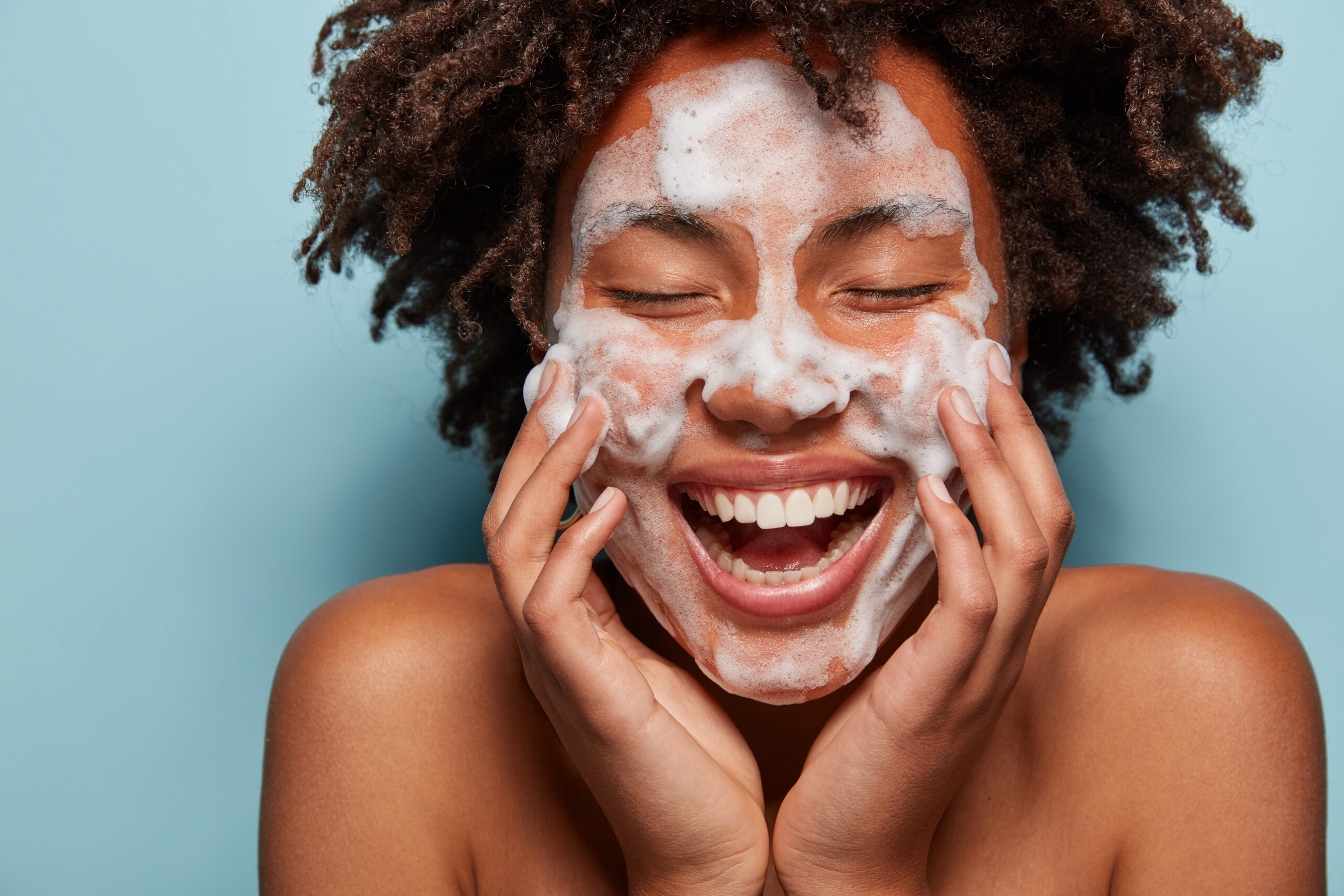 People, skin care and rejuvenation concept. Pleased dark skinned woman with soft skin, applies white foam on face, touches cheeks, smiles broadly, cares of hygiene, feels relaxed and satisfied. Face wash cleanser