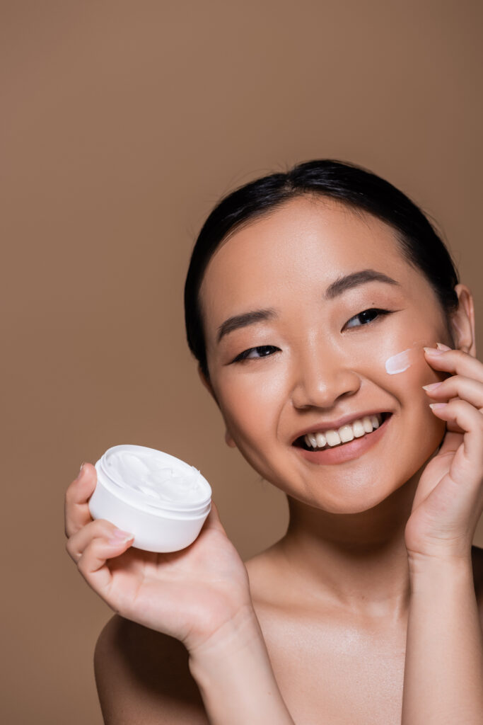 Positive Asian woman applying face moisturizer isolated on brown. cream face. face moisturizer. skincare. face care