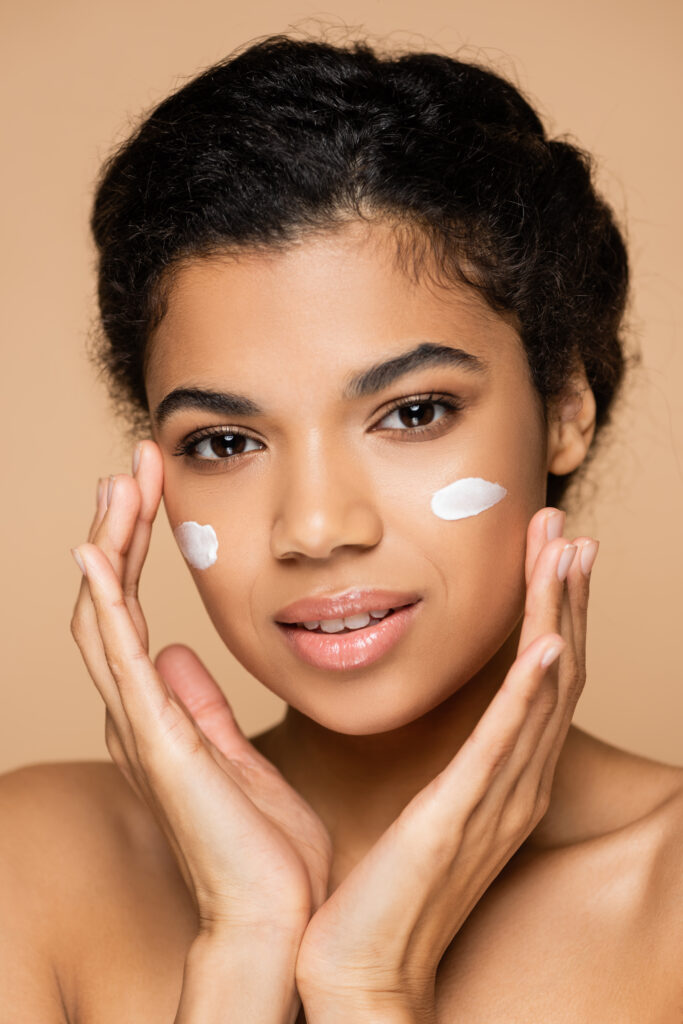 young African American woman with face cream on cheeks isolated on beige. face moisturizer. face care. skincare