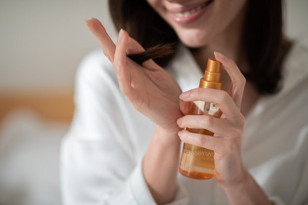 Cheerful young european woman applies spray hair serum in bedroom interior, close up, cropped, blurred. Beauty care, treatment of dry hair and split ends at home, cosmetics for procedures