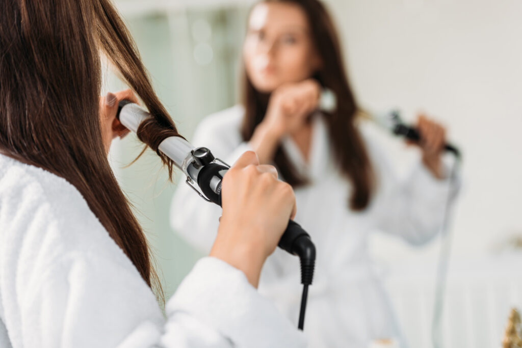 Woman Styling Hair with Curling Iron for Curly or Wavy Look. Curly hair. Curling irons. cropped shot of brunette girl in bathrobe using hair curler at mirror in bathroom.