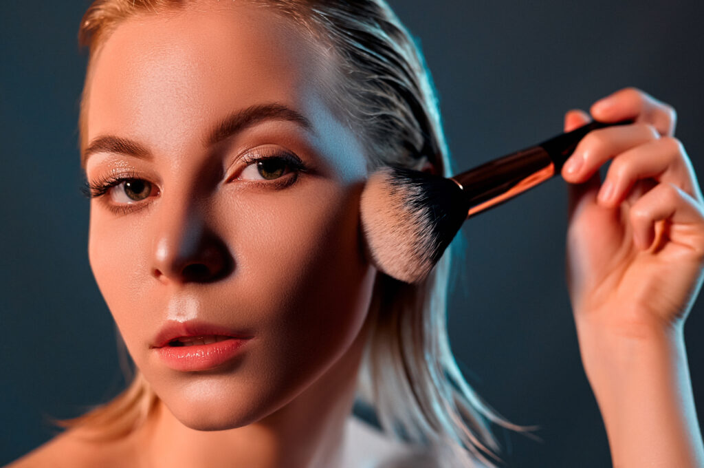 Face Highlighters application tips and technique. A beautiful girl with blonde hair looks at the camera and puts makeup on her face. Gray background. Beauty concept.