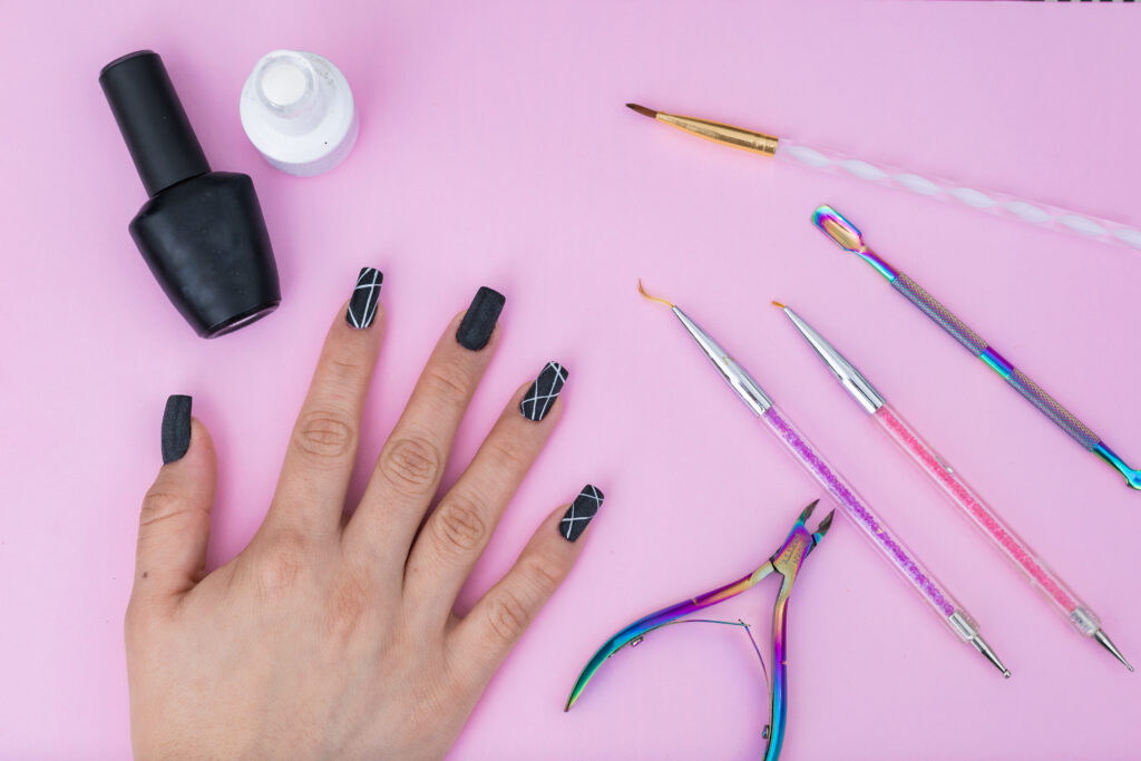 hand of a Latin girl, surrounded by basic elements for a manicure, placed on a pink background. black and white nail polish, colored brushes, cuticle pusher and cuticle nippers. sell of articles. home nail art tools. nail design tools. 