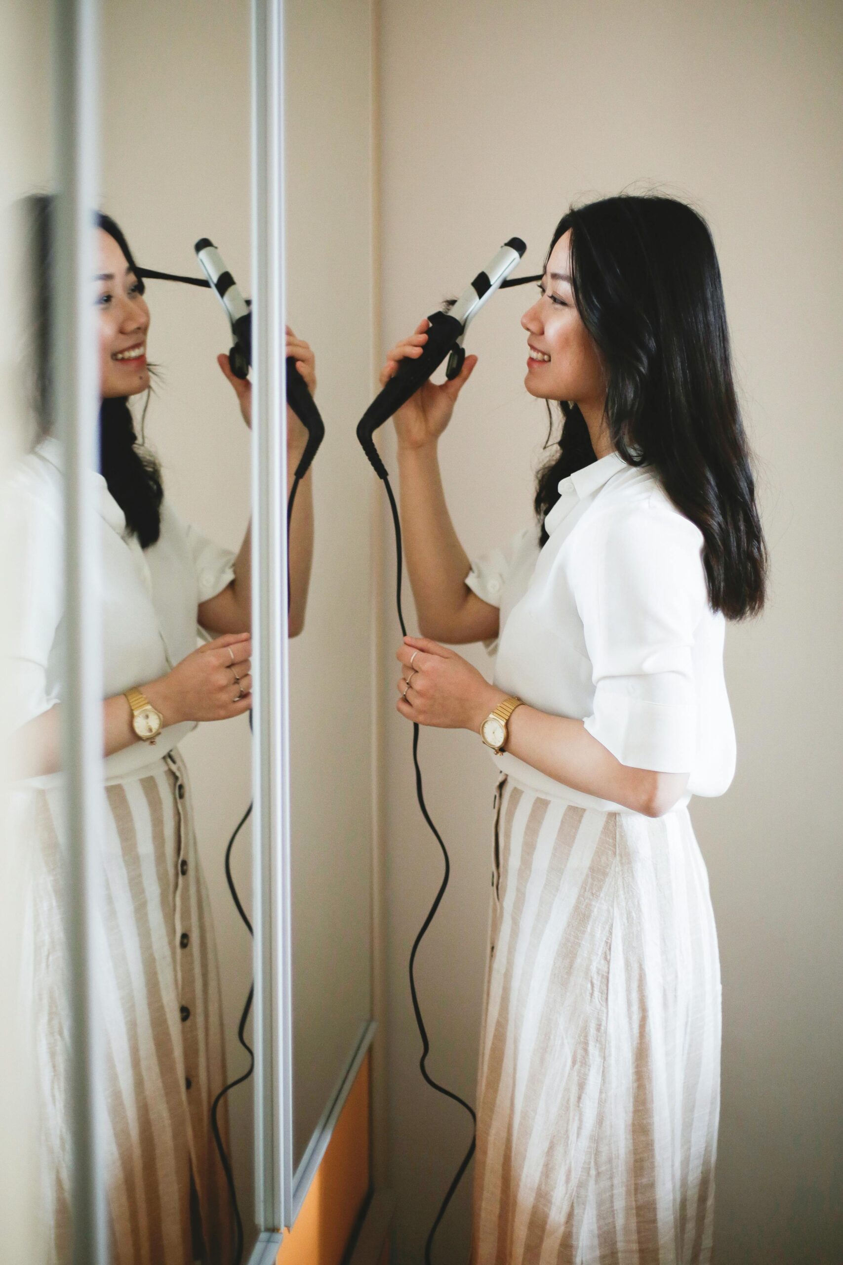 A radiant woman smiles as she styles her hair into waves and curls using a curling iron in front of a mirror.