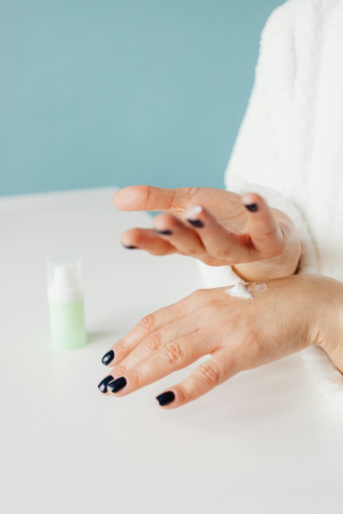 A close-up of a person's hands with glossy, dark nail polish applying lotion, demonstrating essential nail care. The person wears a white robe, suggesting a self-care routine. The light green bottle of lotion on the table complements the scene, illustrating tips for maintaining long-lasting summer nails through regular moisturizing and proper care.