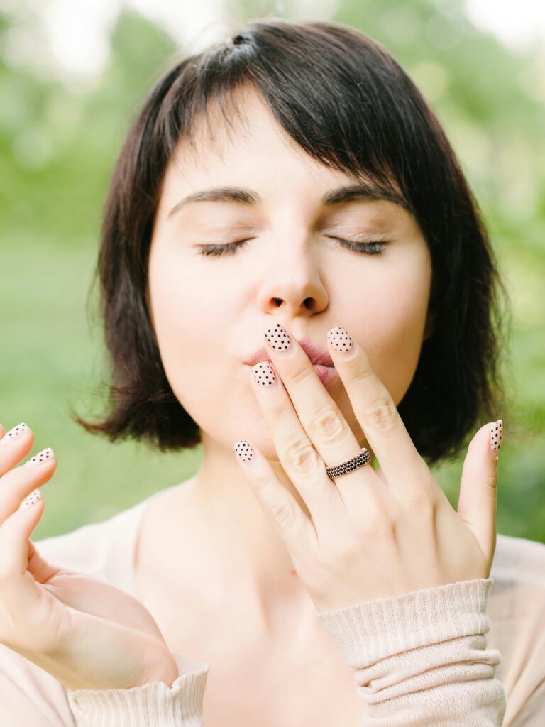 A woman with short, dark hair and closed eyes is gently kissing her hand, showcasing her stylish summer nails. The nails feature a soft nude base with black polka dots, perfectly matching her understated outfit with light beige sleeves. The look is complemented by a textured ring, creating a harmonious and chic summer style.