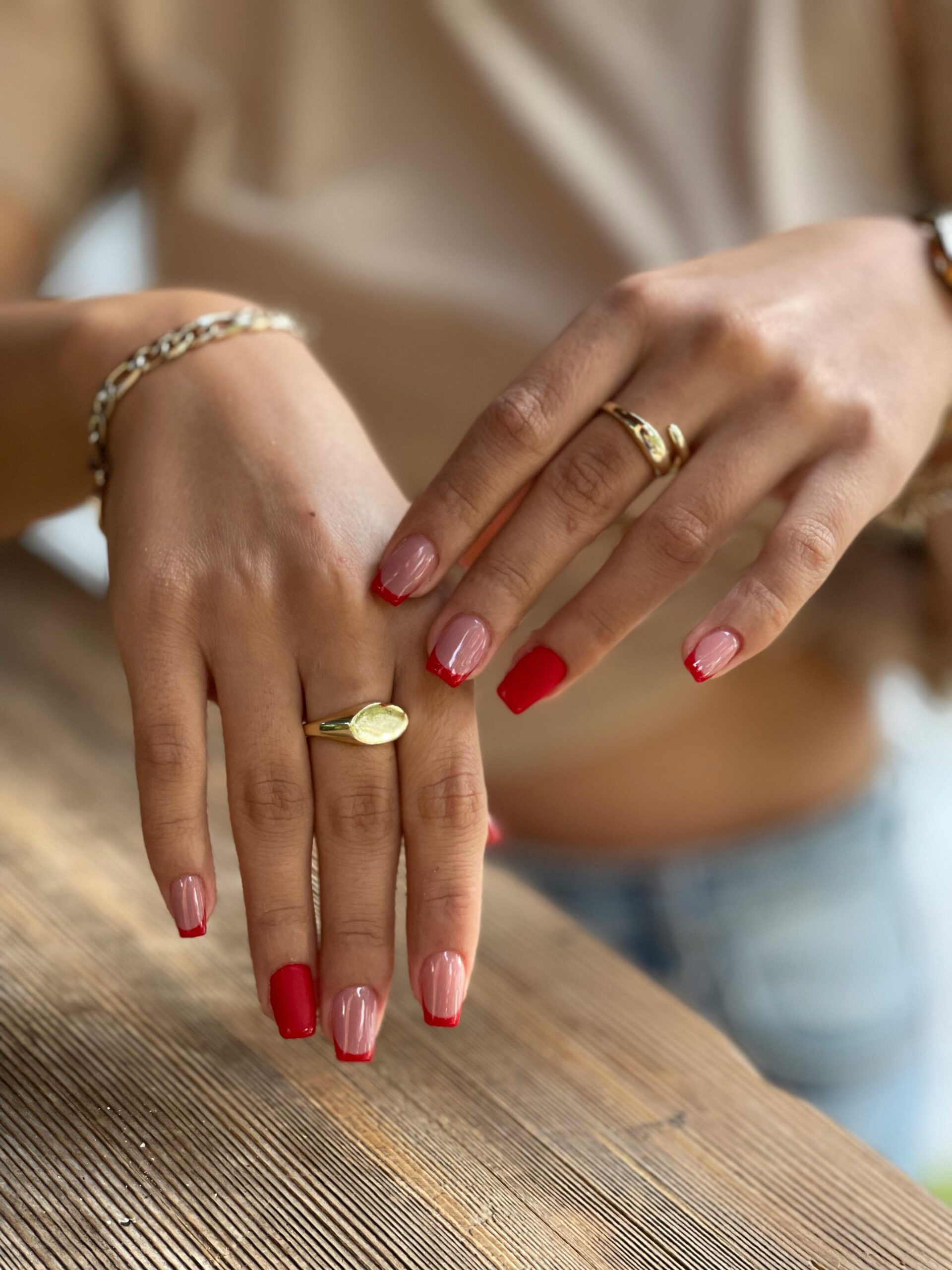 A faceless woman's nails, adorned with gold rings, apply an eye-catching red summer nail polish scented with a hint of pink. Summer 2024 nail polish shades