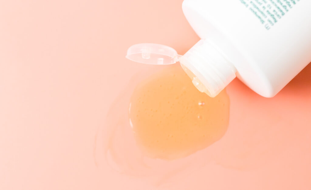 One white bottle with shampoo flowing out of it lies on a peach background with a small copy space, flat lay closeup. The concept of cosmetics, isolation, personal hygiene. face care