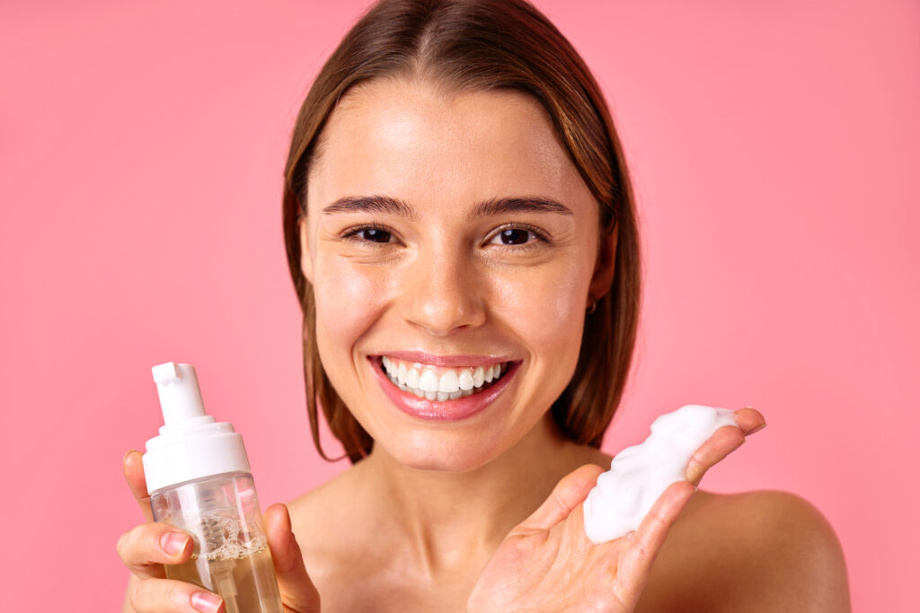 Beautiful woman cleansing her face with foam isolated on pink background. Facial skin care, cosmetology, beauty concept.